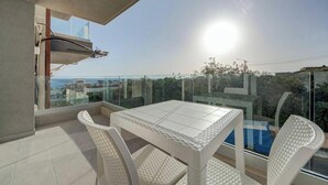 Uma varanda com mesa e cadeiras, guarda-corpo de vidro e vista para a cidade.