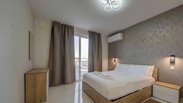 Quarto moderno com cama de madeira, mesa de cabeceira, pequeno armário de madeira e vista para o exterior.