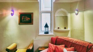 Una habitación con ventana, radiador, sofá rojo, una mesita y un cuadro enmarcado en la pared.