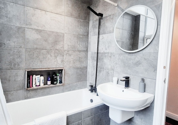 A bathroom with a bathtub, a sink, a mirror, and a shelf with toiletries.
