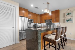 A kitchen with wooden cabinets, stainless steel appliances, a granite island, and wicker bar stools. - Ocean Club.  Gulf Views.  Pool.  Walk to Beach (Biloxi)