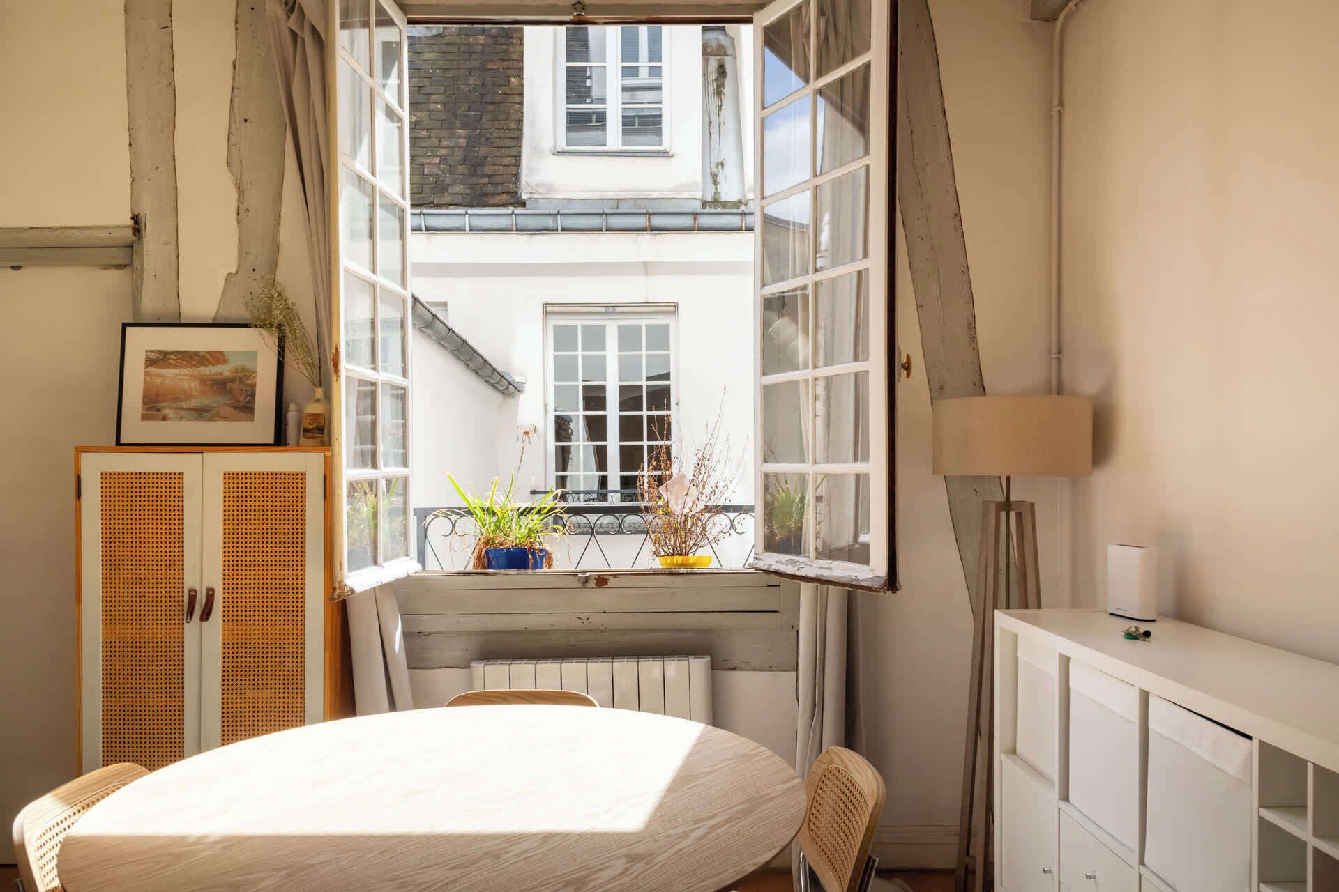 Une pièce avec une grande fenêtre ouverte, une table ronde, une chaise en osier, un radiateur et un meuble.