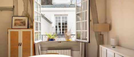 Une pièce avec une grande fenêtre ouverte, une table ronde, une chaise en osier, un radiateur et un meuble.