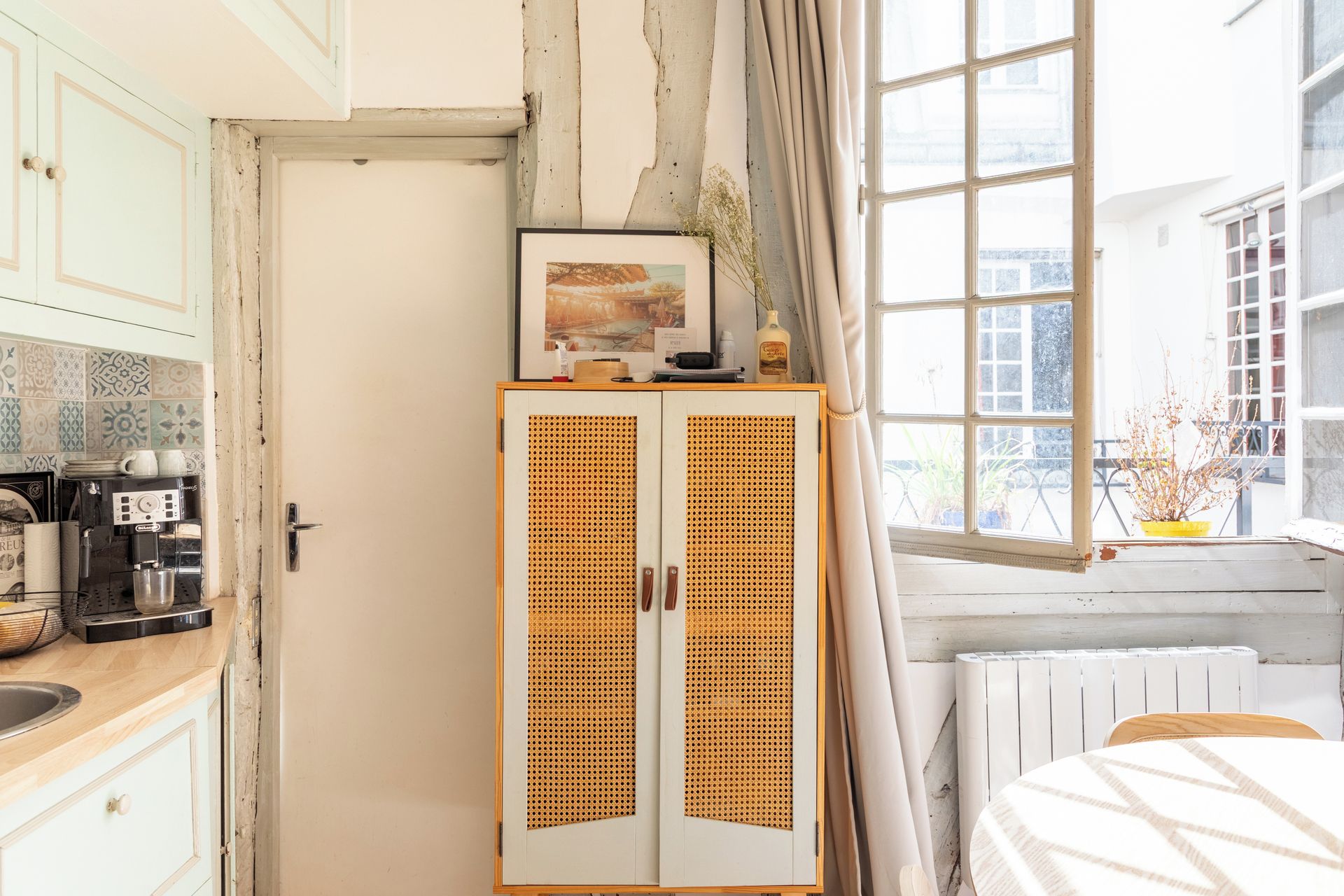 A room with a kitchenette, a rattan cabinet, a window with curtains, and a radiator.
