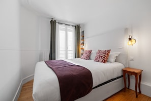 Ein Schlafzimmer mit Bett, Nachttisch, einem Fenster mit Vorhängen und einer Wandleuchte.