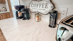 A kitchen with marble countertops, a coffee sign, a coffee maker, a toaster, and wooden cabinets. - Emily’s Beach Shack at Holden Beach! Charming NEW home about 1 mile from beach! (Supply)