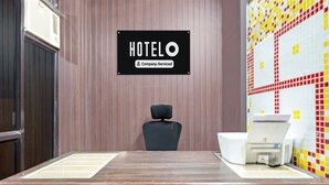 A hotel room with a wooden panel wall, a sign indicating it's a company-serviced hotel, a black chair, a desk with a printer, and a tiled wall.