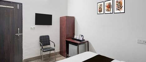 A hotel room with a bed, a desk with a coffee maker, a chair, a TV mounted on the wall, and framed artwork.