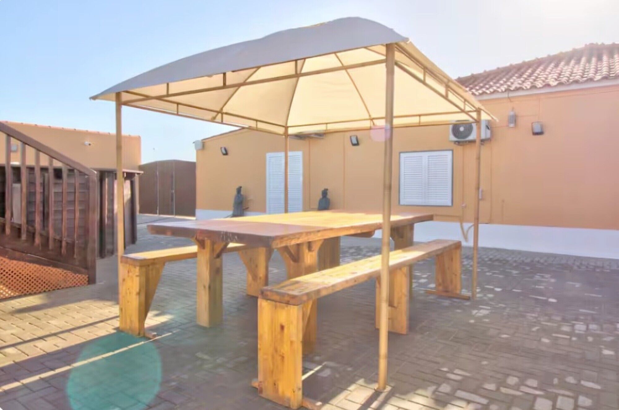 Uma mesa de madeira ao ar livre com bancos sob um toldo, um edifício com telhado em cerâmica ao fundo.