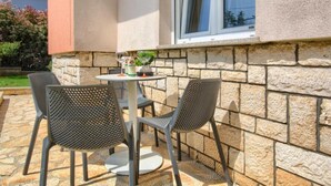 Eine Terrasse im Freien mit einem runden Tisch, zwei Stühlen, einem Topfpflanzen und einem Fenster mit Vorhängen.