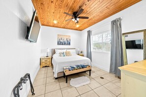 Ein Schlafzimmer mit Holzboden, einem Bett mit weißer Bettdecke, einem Nachttisch, einem Beistelltisch, einem Fenster mit Vorhängen und einem an der Wand montierten Fernseher.