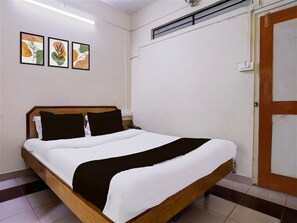 A single bed with a wooden frame, white bedding, and two dark pillows. There are three framed artworks on the wall, a door with a wooden frame, and a tiled floor. - Deluxe Brookefield Central Bangalore Inn (Bengaluru)