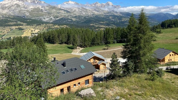A wooden cabin with a sloped roof, surrounded by a forested area and mountains in the background. - 4 bed apartment - La Joue du Loup (Dévoluy)