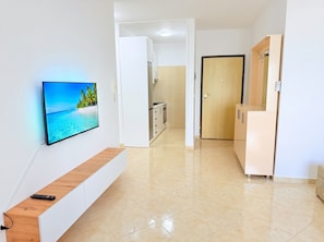 A modern hotel room with a TV on the wall, a wooden bench, a kitchen area, and a wooden door.