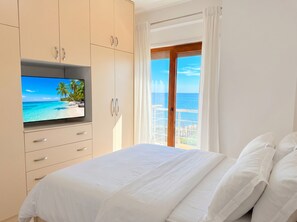 A bedroom with a TV displaying a beach scene, a balcony with a view of the sea, and a bed with white bedding. - Sea View Apartment In Durres with Private Parking (Durres)