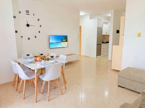 A dining area with a clock, a TV displaying a beach scene, and a kitchen in the background.