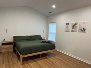 A bedroom with a wooden bed, a nightstand, a wall-mounted light, a window with blinds, and three framed artworks. - “Stylish 3BR Near NoDa w/ Outdoor Lounge" (Charlotte)