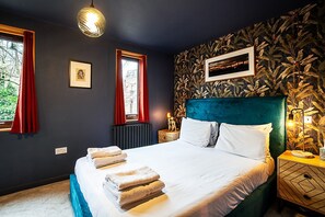 Ein Schlafzimmer mit einem Bett, zwei Fenstern mit roten Vorhängen, einem Heizkörper, einer Wand mit einem Blattmuster und einem Nachttisch mit einer Lampe.