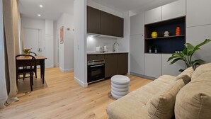 Ett modernt kök med inbyggda vitvaror, en matplats med ett träbord och stolar samt ett vardagsrum med en beige soffa och en pall.