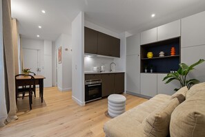Eine moderne Küche mit integrierten Geräten, ein Essbereich mit Holztisch und -stühlen sowie ein Wohnzimmer mit beiger Couch und Hocker.