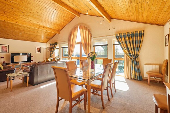 A spacious living area with a high wooden ceiling, a glass dining table surrounded by chairs, a sofa, and a television.