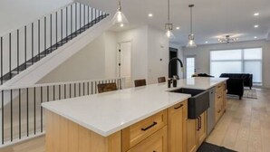 A modern kitchen with a central island, wooden cabinets, and a staircase with a glass railing. - Chic Retreat 4 BR Near UofC & HWY , Free Parking (Calgary)