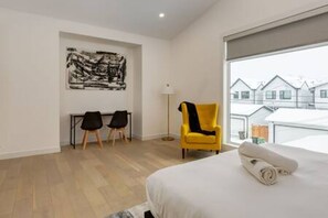 Habitación de hotel con una cama, un sillón amarillo, un escritorio con dos sillas, una lámpara de pie y un ventanal con vistas a casas.