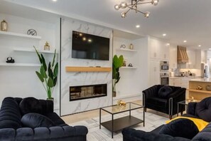 Una sala de estar moderna con chimenea, sofás negros, una mesa de centro de vidrio y un televisor montado en la pared.