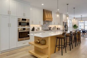 A modern kitchen with white cabinetry, stainless steel appliances, a large island with bar stools, and pendant lighting. - Chic Retreat 4 BR Near UofC & HWY , Free Parking (Calgary)