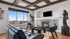 A living room with a leather sofa, a fireplace, a TV mounted on the wall, and a large window. - "Amantara" 5 BR Luxury Retreat | Bright & Serene (Calgary)