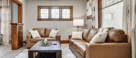 A living room with a brown leather sofa, a wooden coffee table, and a large window with blinds.