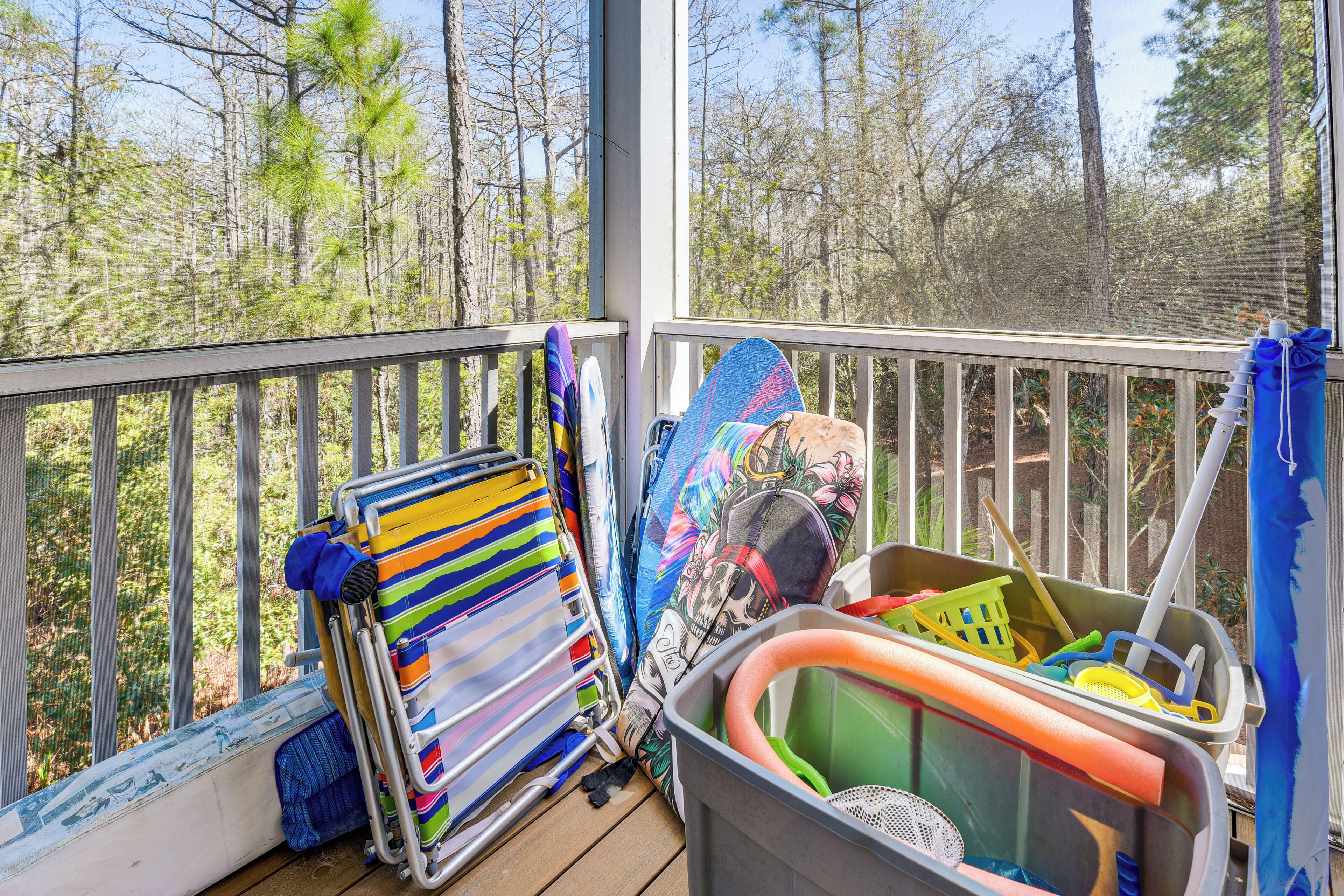 A balcony with a view of trees, a beach chair, a surfboard, and a laundry basket.