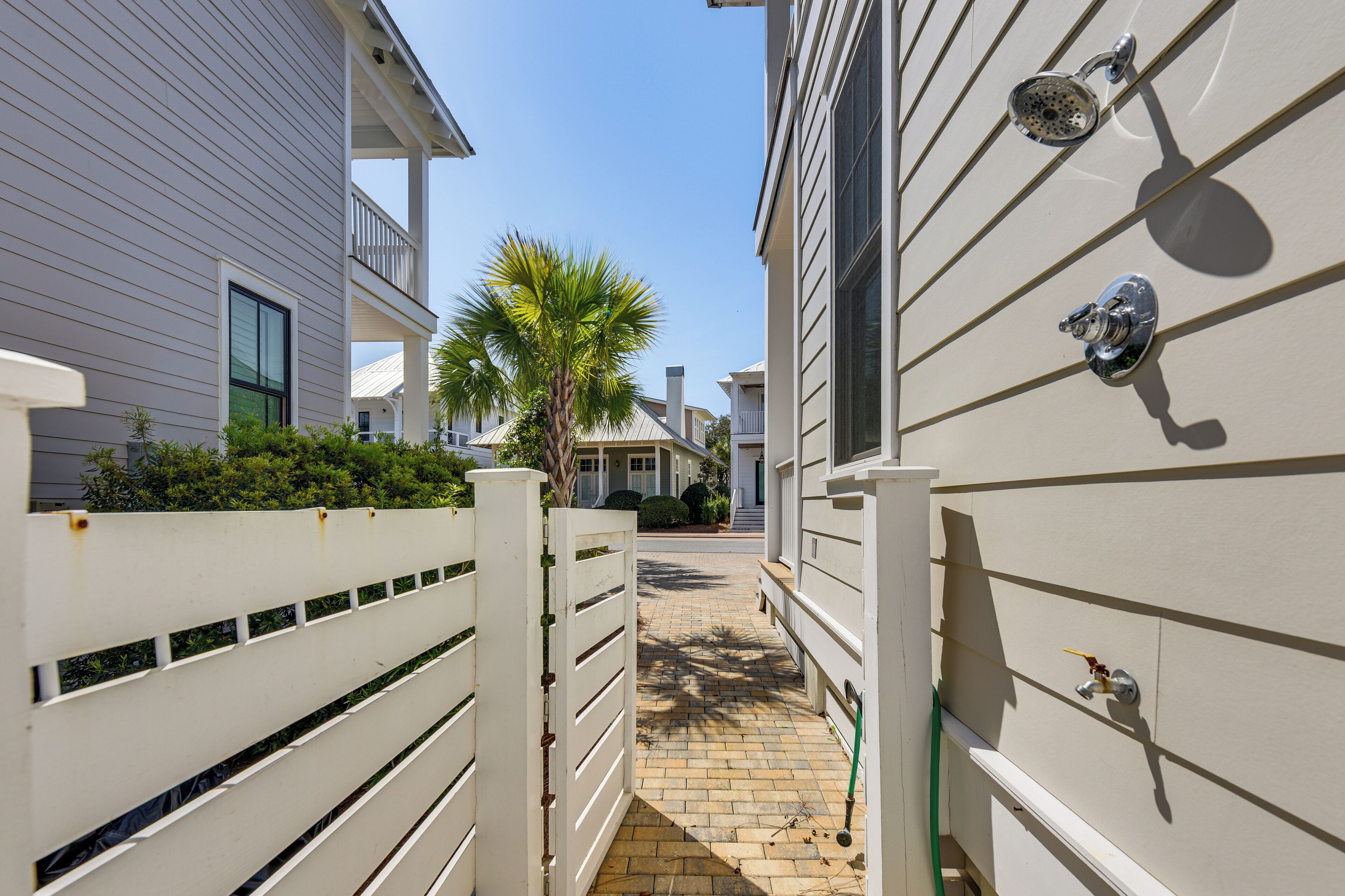A narrow pathway between two buildings with a palm tree, a white fence, and a metal gate.