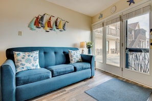 A blue sofa with patterned pillows, a wall-mounted sailboat decoration, and a view of a building through a large window. - Steps to Sand! Oceanfront Gem in Old Orchard Beach (Old Orchard Beach)
