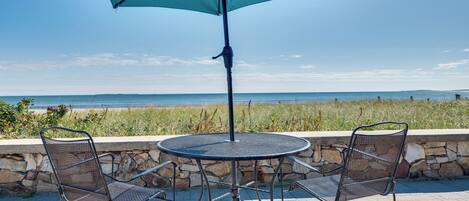A round metal table with two chairs and a large blue umbrella, set against a stone wall with a view of the sea.