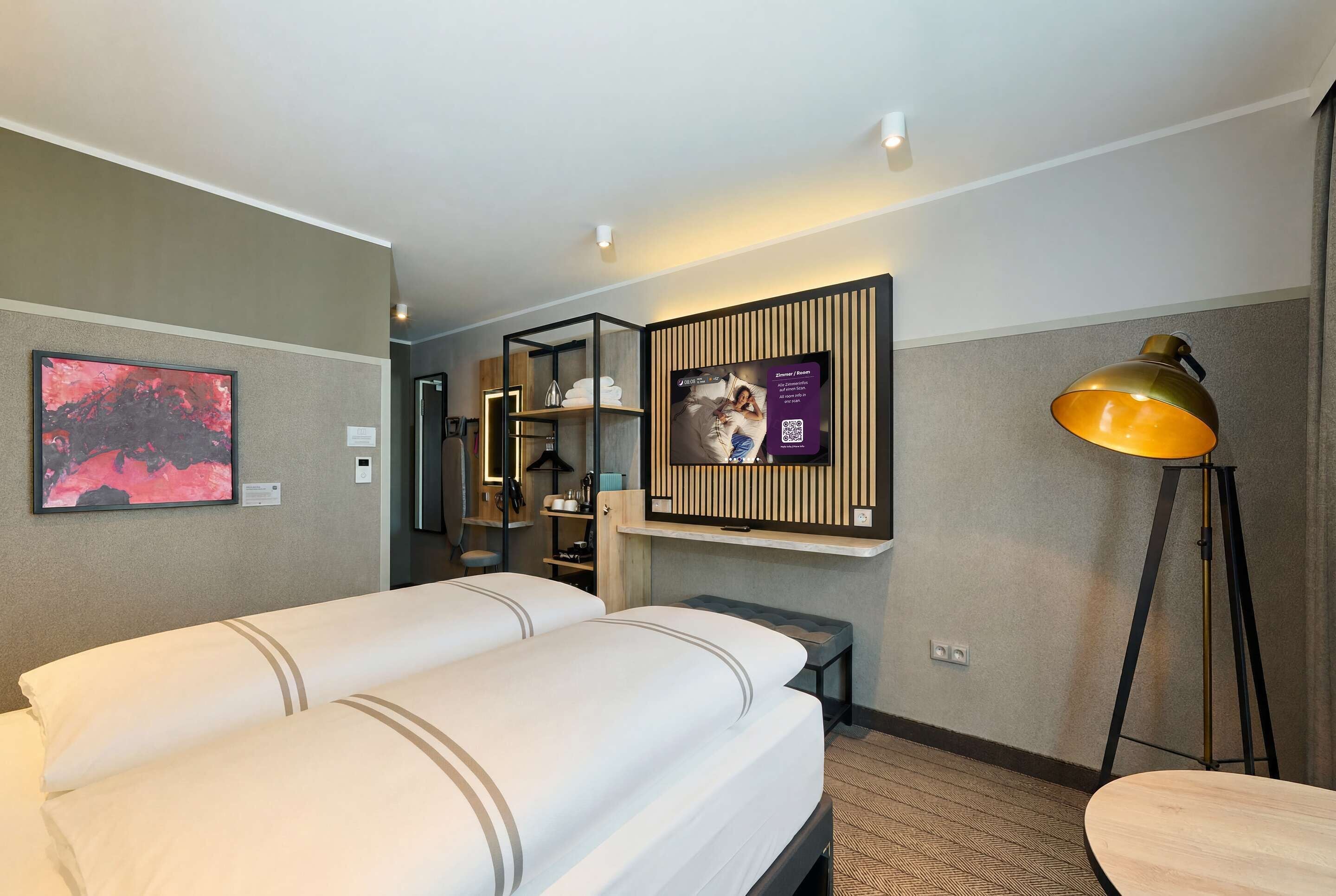 Une chambre d’hôtel avec deux lits, un bureau, une lampe, une télévision et une étagère fixée au mur.