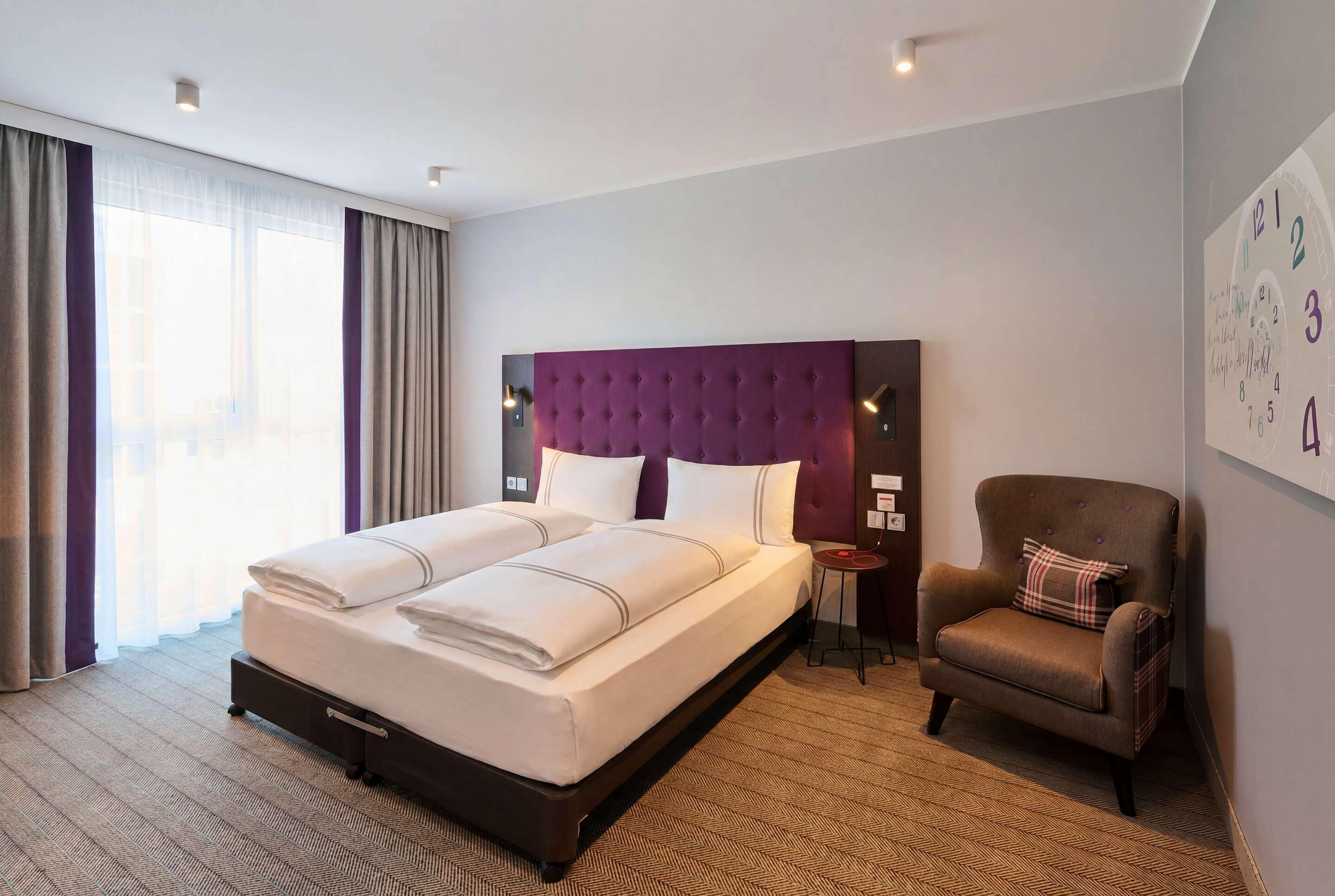 Habitación de hotel con una cama grande, una silla, una mesita y un reloj de pared.