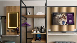 A hotel room with a wooden door, a mirror, a hairdryer, a shelf with towels, a coffee maker, a TV displaying room information, and a trash bin. - Premier Inn Frankfurt Eschborn (Eschborn)