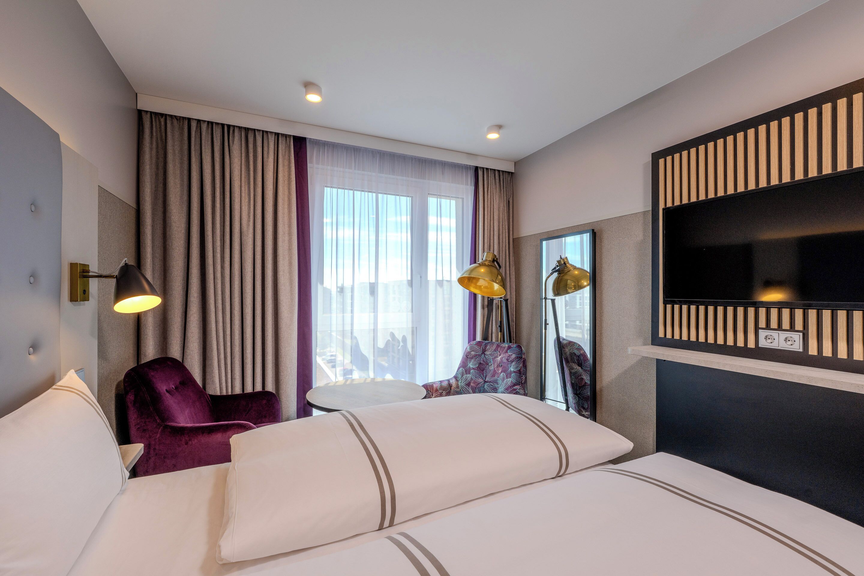A hotel room with a bed, a chair, a small table, a TV, and a window with curtains.