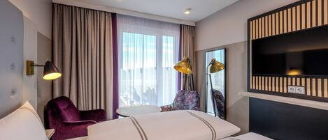 A hotel room with a bed, a chair, a small table, a TV, and a window with curtains.