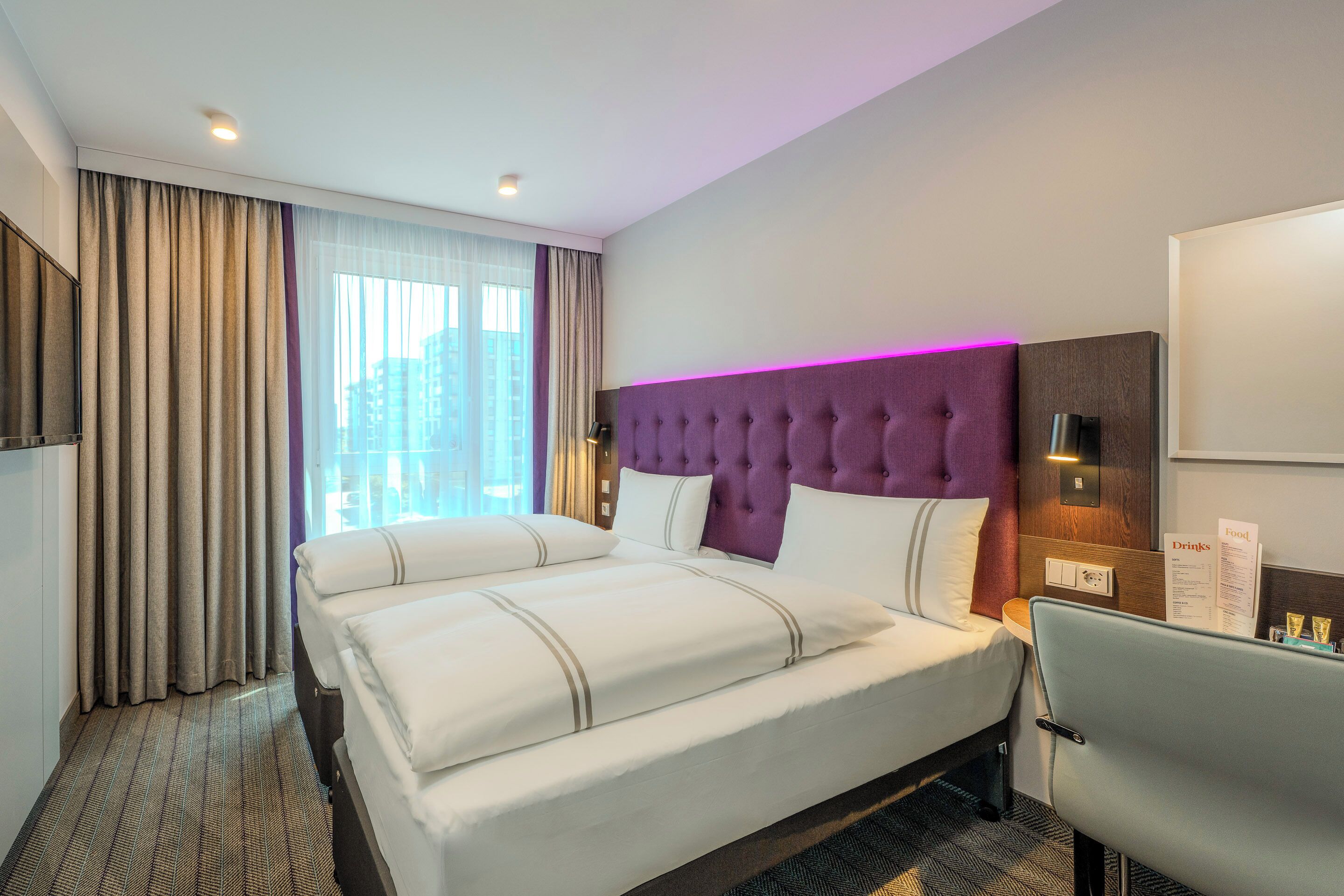 Habitación de hotel con una cama grande, un escritorio, una silla, cabecera morada y una ventana con cortinas.