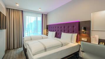 Habitación de hotel con una cama grande, un escritorio, una silla, cabecera morada y una ventana con cortinas.