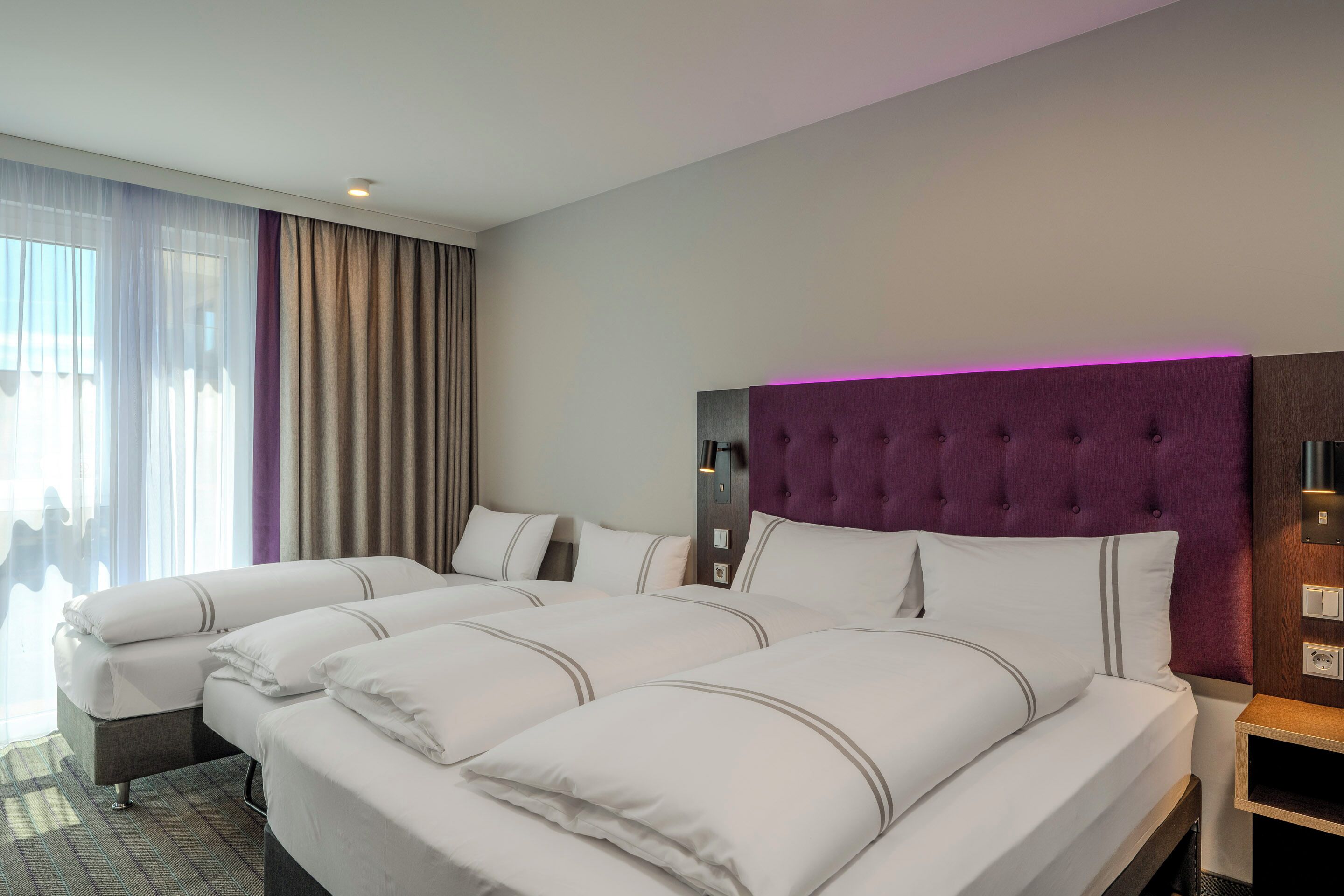 A hotel room with two beds, a large window with curtains, a purple headboard, and a bedside table.