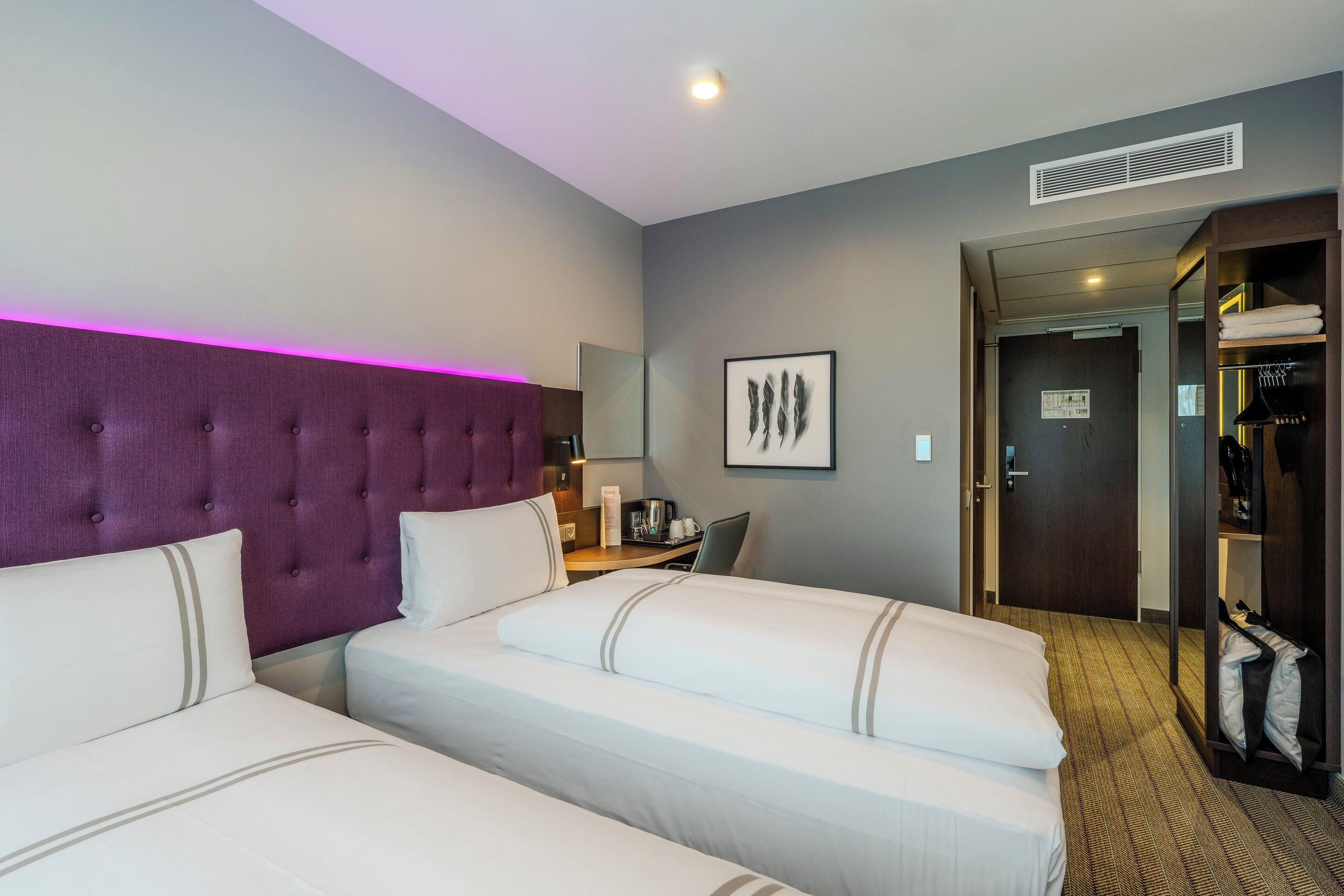 Ein Hotelzimmer mit zwei Betten, einem violetten Kopfteil, einem Schreibtisch mit Stuhl, einem Spiegel und einem Kleiderschrank.
