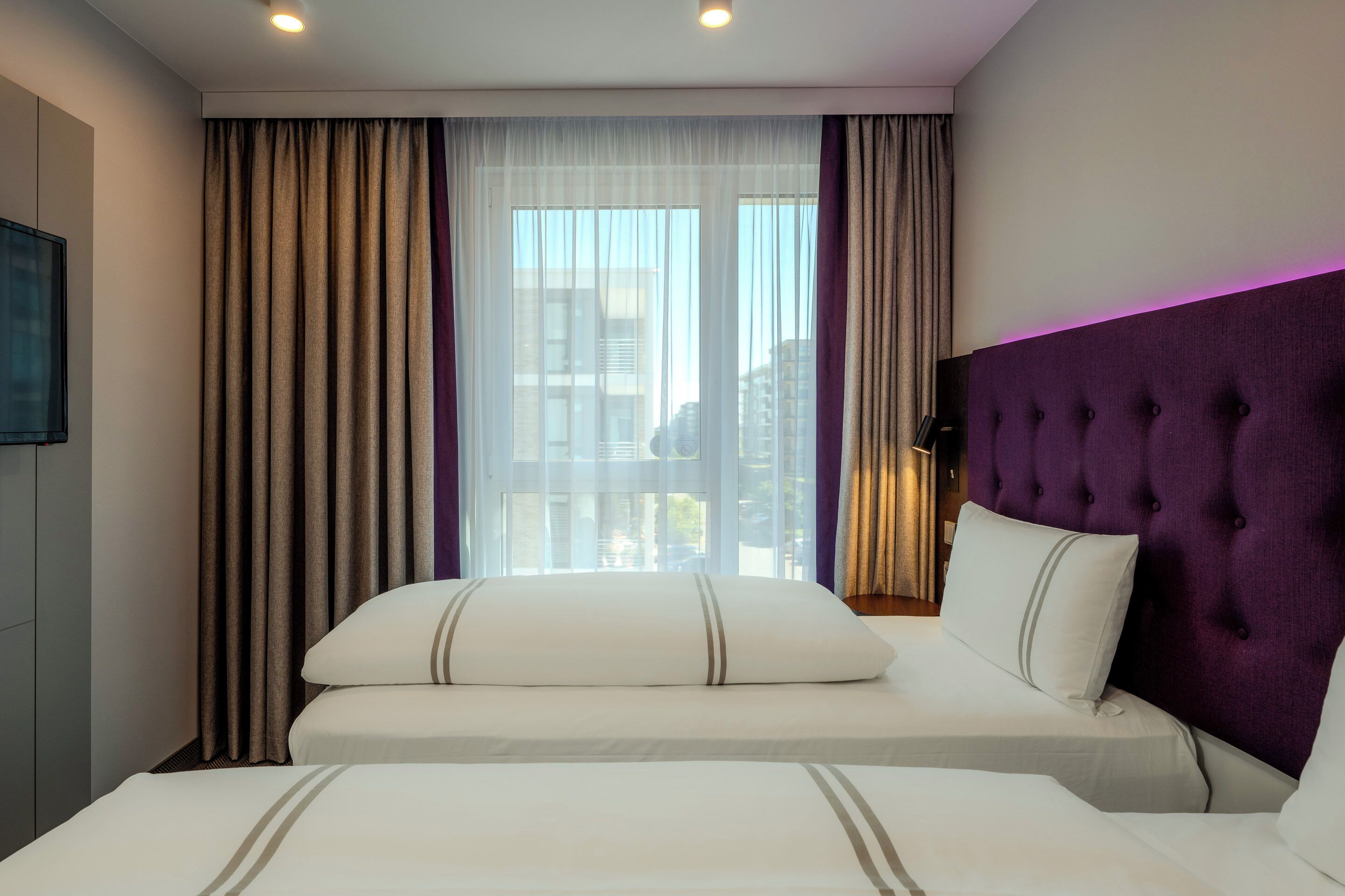 Une chambre d’hôtel avec deux lits, une grande fenêtre avec des rideaux, une tête de lit violette et un téléviseur à écran plat fixé au mur.