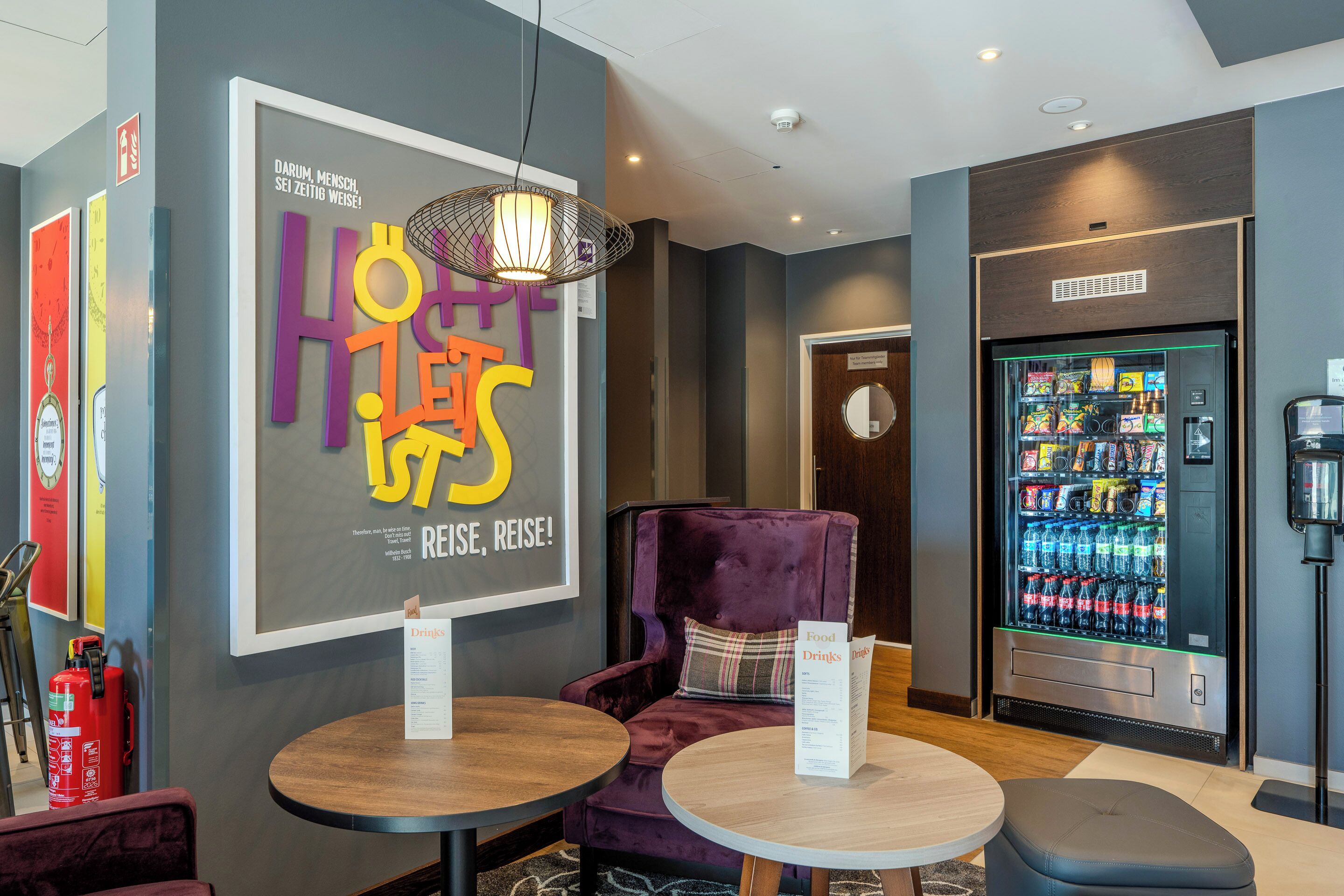 A hotel lobby with a large sign, a vending machine, a purple chair, and a table with a menu.