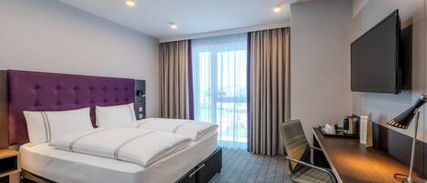 A hotel room with a large bed, a desk with a chair, a flat-screen TV, and a window with curtains.
