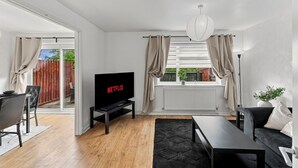 Een moderne woonkamer met een televisie waarop 'NETFLIX' staat, een eethoek met een tafel en stoelen, en uitzicht op een houten terras door glazen deuren.