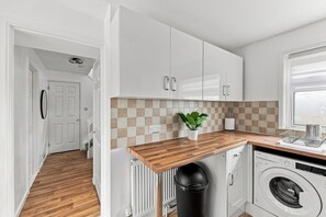 Cuina compacta amb armaris blancs, taulell de fusta, paperera negra, rentadora i una planta en test.