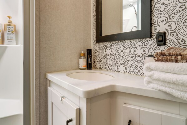 Un baño con lavamanos blanco, un espejo, papel tapiz estampado, un perchero y una repisa con artículos de aseo.